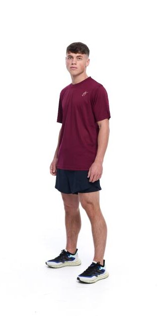 Male wearing red training top and black shorts standing in front of a white background