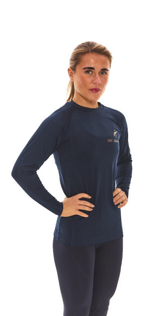 Blonde-haired female model wearing a blue sports base layer, matching blue leggings, and white trainers, standing in front of a white wall with hands on her hips.