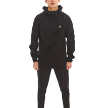 Male model with black hair wearing a black rain jacket and black tracksuit bottoms, standing against a white wall.