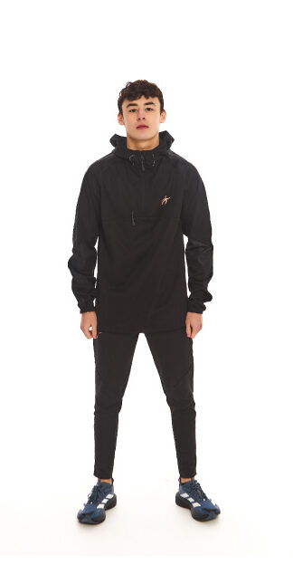 Male model with black hair wearing a black rain jacket and black tracksuit bottoms, standing against a white wall.