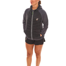Brown-haired woman in black athletic quarter-zip and black shorts standing against a white wall.