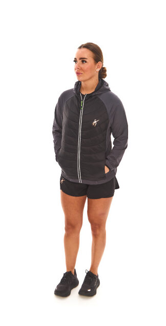 Brown-haired woman in black athletic quarter-zip and black shorts standing against a white wall.