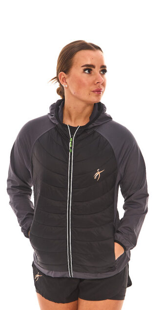 Brown-haired woman in black athletic quarter-zip and black shorts standing against a white wall.