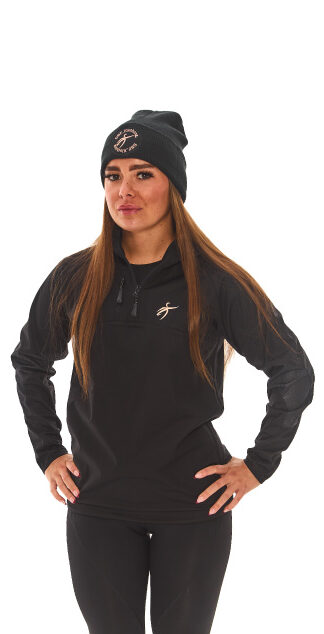 Brown-haired woman in black athletic tracksuit top and base layer bottoms standing against a white wall with hands on hips.