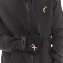 Male model wearing a black sports woolen hat and black rain jacket, looking down at the jacket logo while standing against a white wall.