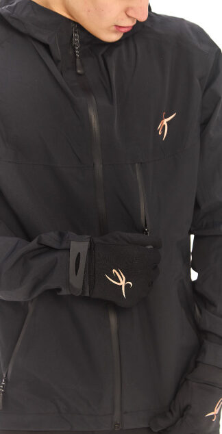 Male model wearing a black sports woolen hat and black rain jacket, looking down at the jacket logo while standing against a white wall.