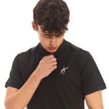 Male model with black hair wearing a black polo shirt and black tracksuit bottoms, standing against a white wall and looking at the logo on his shirt.