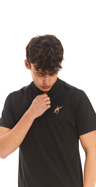 Male model with black hair wearing a black polo shirt and black tracksuit bottoms, standing against a white wall and looking at the logo on his shirt.