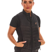 Brown-haired female model wearing a black sports gilet, black leggings, and black trainers, standing against a white wall with hands in pockets.