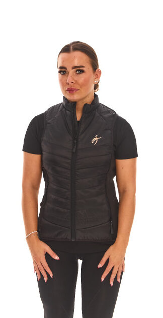 Brown-haired female model wearing a black sports gilet, black leggings, and black trainers, standing against a white wall with hands on hips.