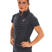 Blonde-haired female model wearing a blue sports gilet, matching blue leggings, and white trainers, standing in front of a white wall with hands on her hips.