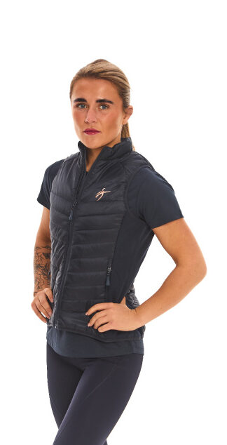 Blonde-haired female model wearing a blue sports gilet, matching blue leggings, and white trainers, standing in front of a white wall with hands on her hips.