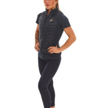 Blonde-haired female model wearing a blue sports gilet, matching blue leggings, and white trainers, standing in front of a white wall with hands on her hips.