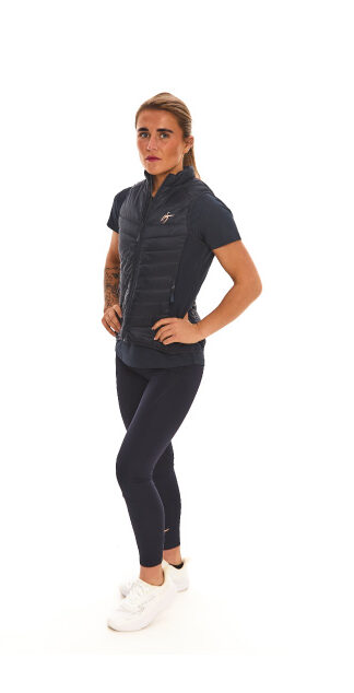 Blonde-haired female model wearing a blue sports gilet, matching blue leggings, and white trainers, standing in front of a white wall with hands on her hips.