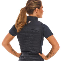 Blonde female model shown from the back, wearing a blue sports gilet and matching leggings, standing against a white wall with hands on hips.