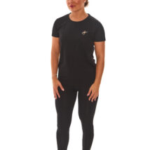 Model with brown hair wearing black sports base layers, black leggings, and black trainers, standing against a white wall with hands on hips.