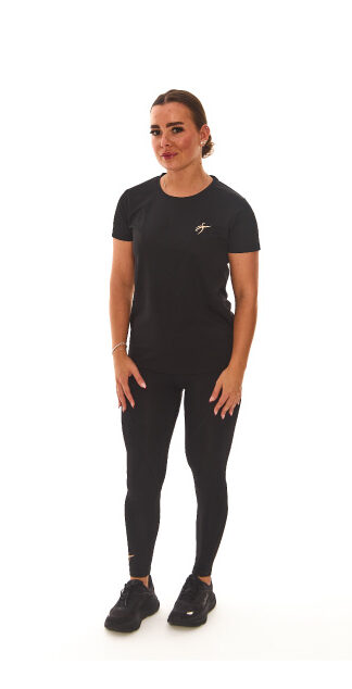 Model with brown hair wearing black sports base layers, black leggings, and black trainers, standing against a white wall with hands on hips.