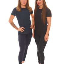 Two female models, one blonde and one brown-haired, wearing black and blue sports base layers, standing in front of a white wall.