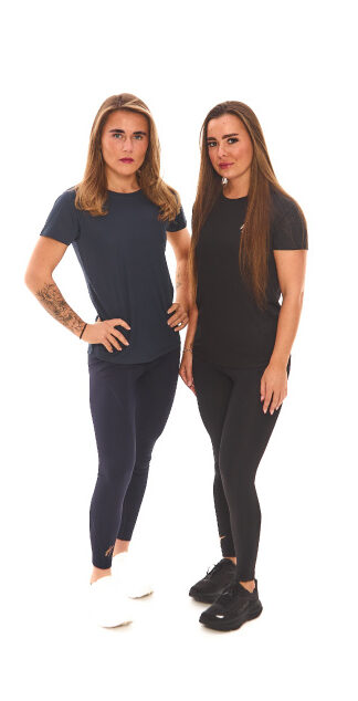 Two female models, one blonde and one brown-haired, wearing black and blue sports base layers, standing in front of a white wall.